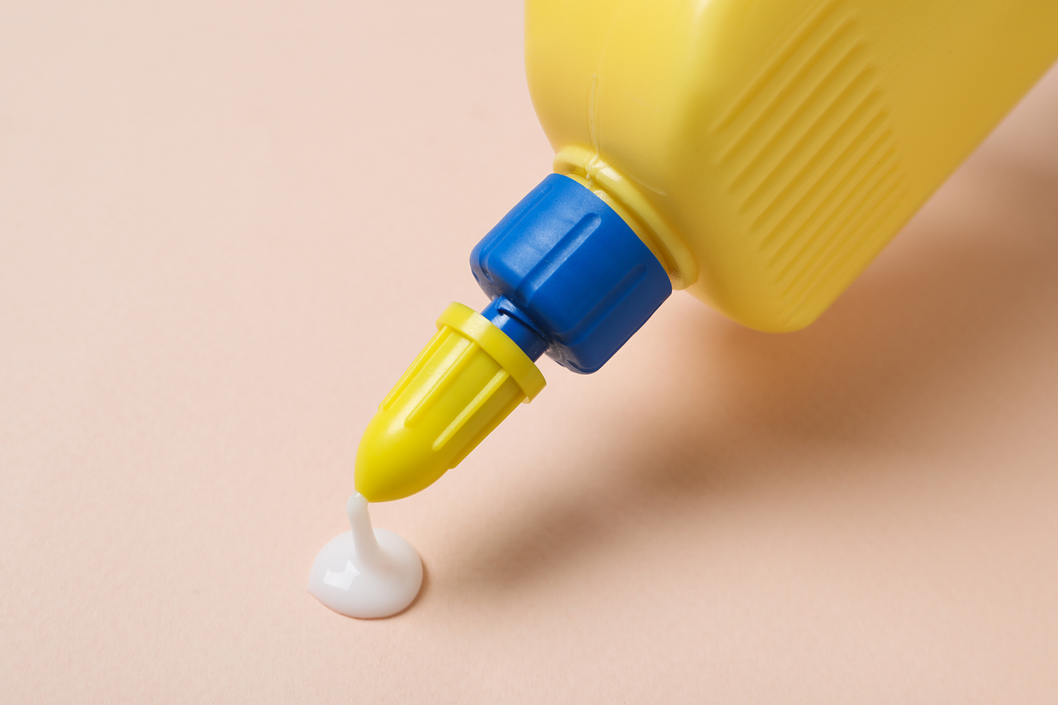 Yellow bottle with blue rim, squeezing out a drop of white PVA glue against a pastel pink background.
